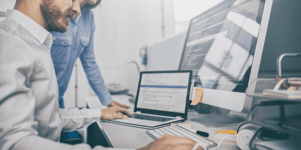 Uma mulher e um homem em frente a uma mesa com ítens de escritório com notebook e um monitor