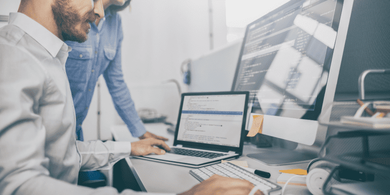 Uma mulher e um homem em frente a uma mesa com ítens de escritório com notebook e um monitor